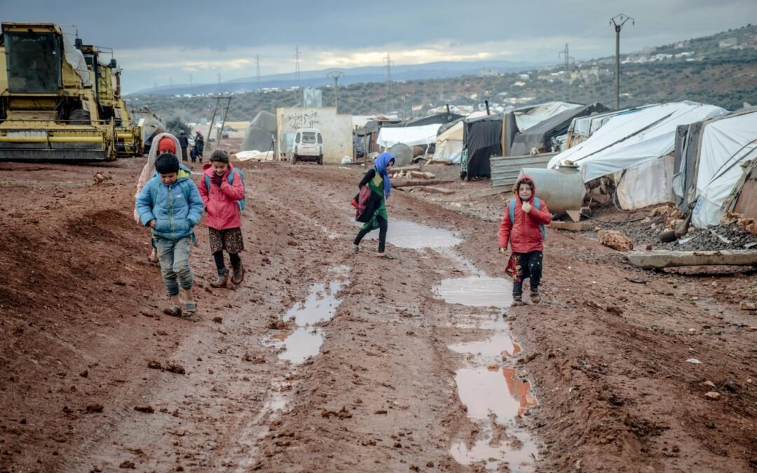 Children walking in muddy conditions near makeshift tents in a humanitarian immigration camp.